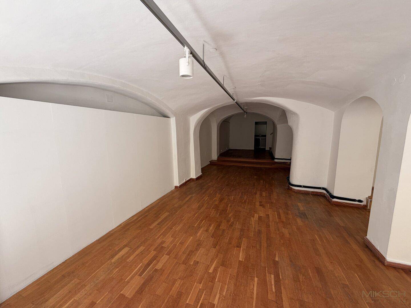 Langer, gewölbter Gewerberaum mit Holzboden und Stufen, die zu einem erhöhten Bereich im Hintergrund führen.