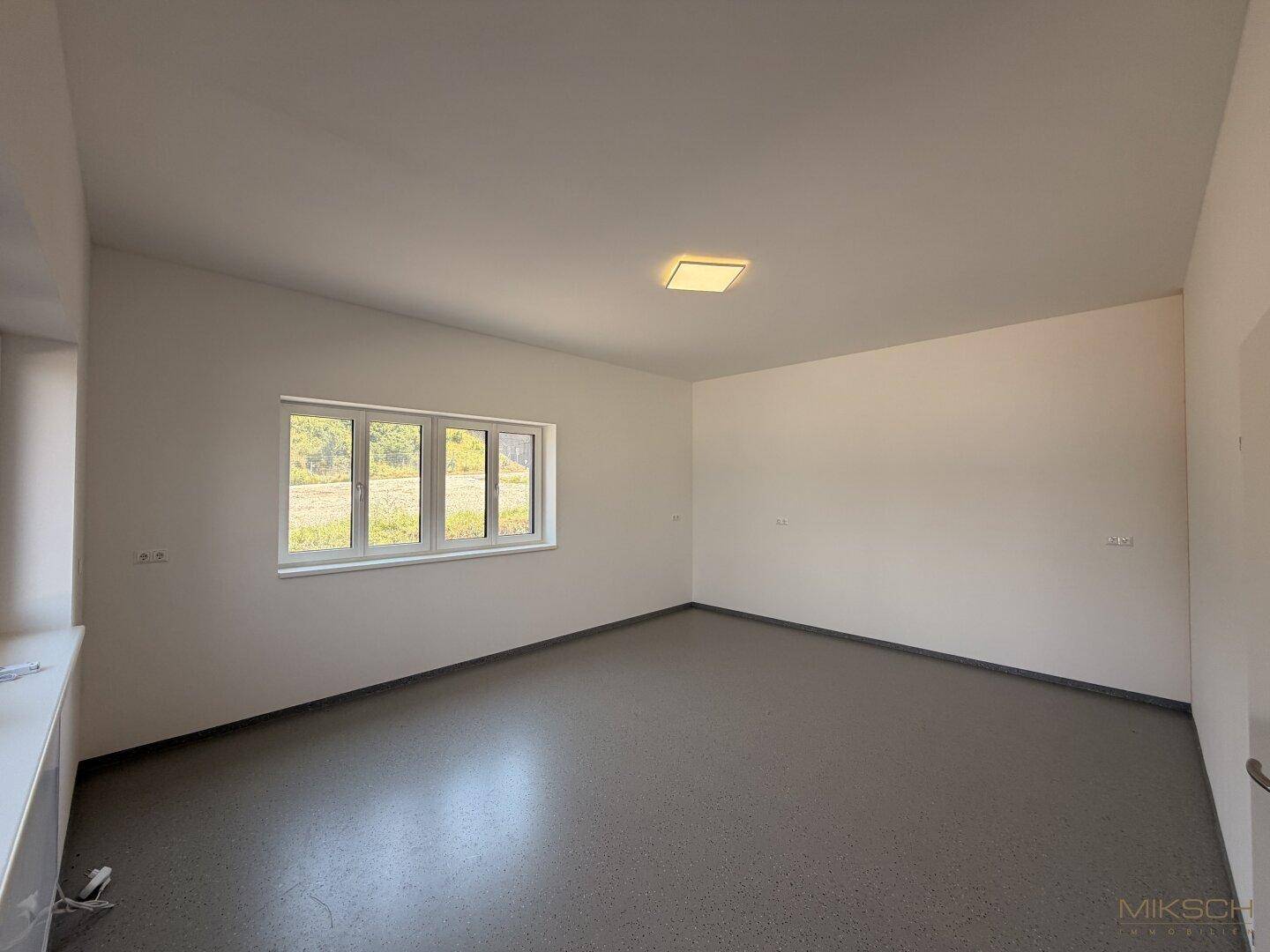 Heller, leerer Raum mit weißen Wänden, grauem Boden und einem Fenster mit Blick ins Grüne.
