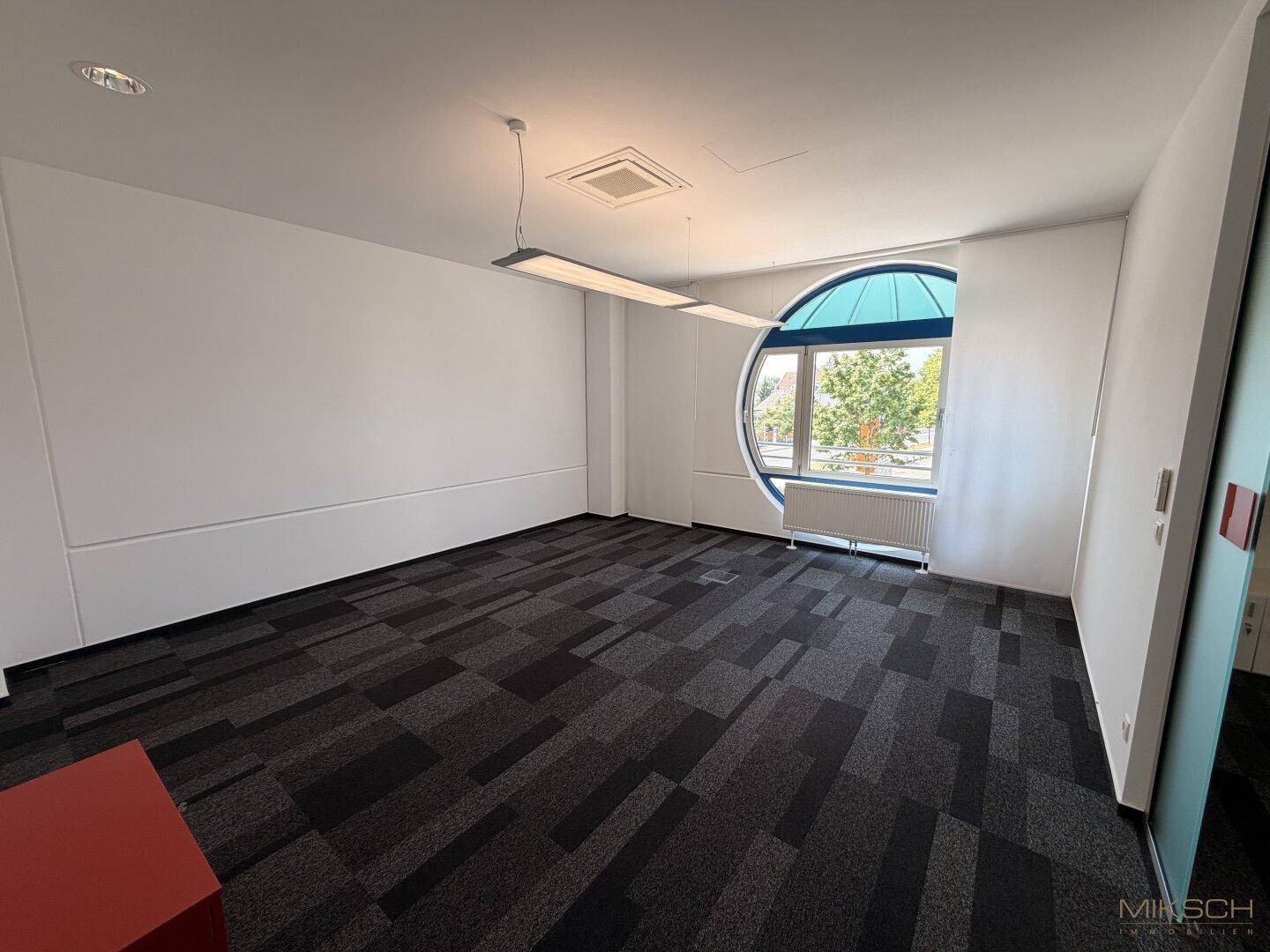 Leeres Büro mit einem markanten runden Fenster, Teppichboden und schlichten weißen Wänden.
