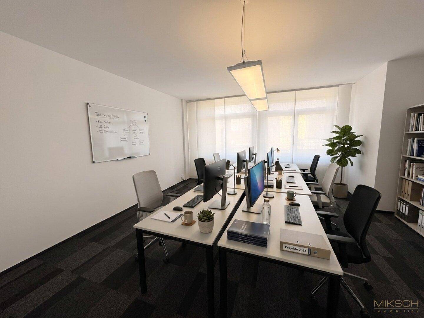 Helles Großraumbüro mit mehreren Arbeitsplätzen, Computern, einem Whiteboard und großen Fenstern.