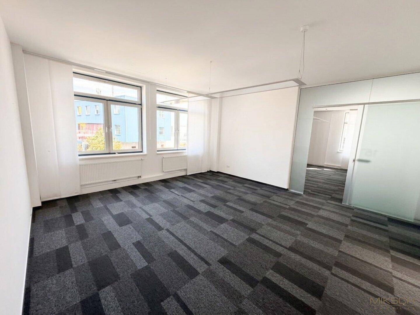 Geräumiges, leeres Büro mit Teppichboden, großen Fenstern und einer Milchglas-Schiebetür.