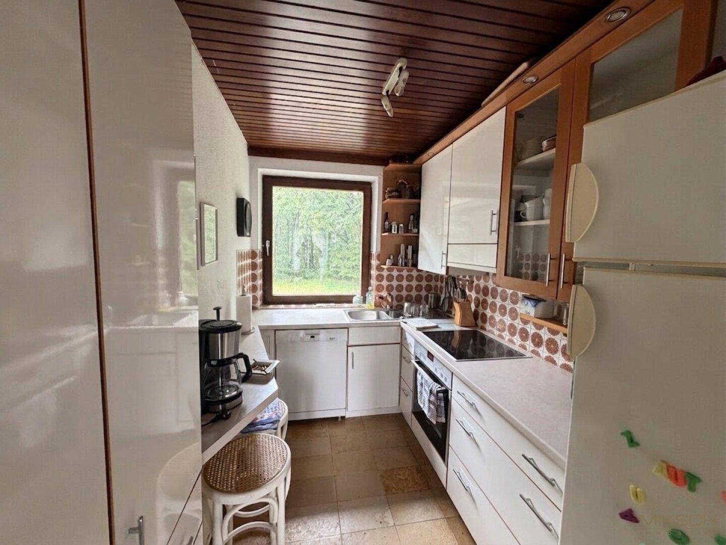 Küche mit weißen Schränken, Holztäfelung an der Decke und Fenster mit Blick ins Grüne.