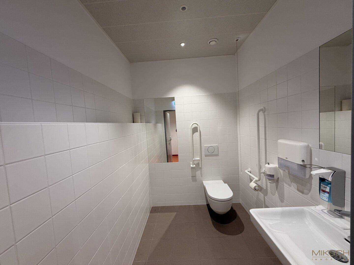 Modernes, barrierefreies Badezimmer mit weißen Fliesen, wandhängendem WC, Waschbecken und Haltegriffen.