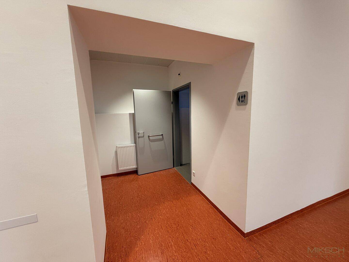 Flurbereich mit Zugang zu den Toiletten, gekennzeichnet durch ein WC-Schild und orangefarbenen Boden.