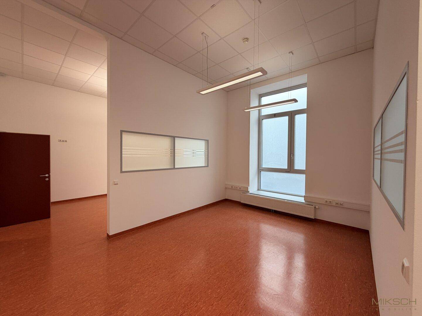 Heller Bürobereich mit einer Trennwand, großen Fenstern und einem durchgehenden Linoleumboden.
