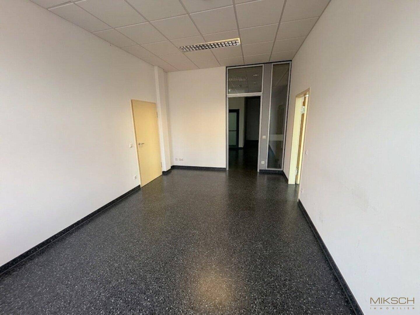 Langer Büroraum mit dunklem Boden und Zugang zu weiteren Bereichen durch Glastüren, bietet flexible Nutzungsmöglichkeiten.