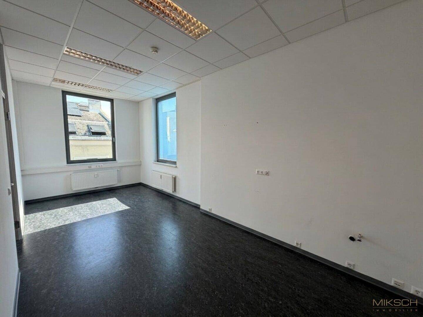 Leeres Büro mit dunklem Boden und zwei Fenstern, die für ausreichend Helligkeit sorgen und einen Stadtblick ermöglichen.