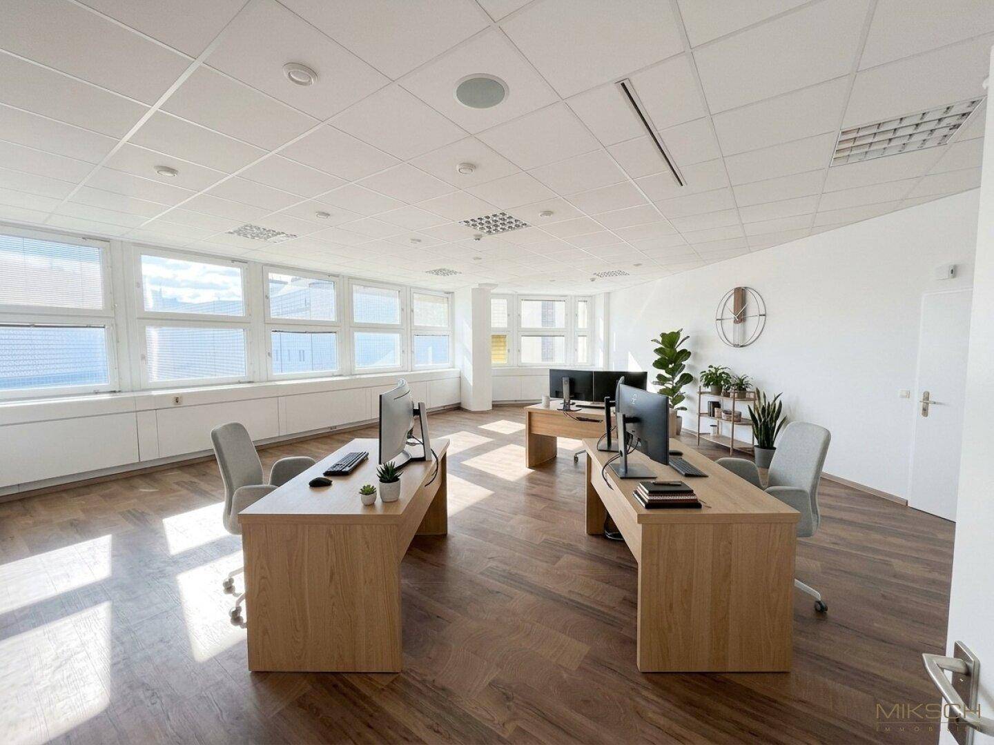 Modern eingerichtetes Büro mit mehreren Arbeitsplätzen, Holzboden und großen Fenstern.
