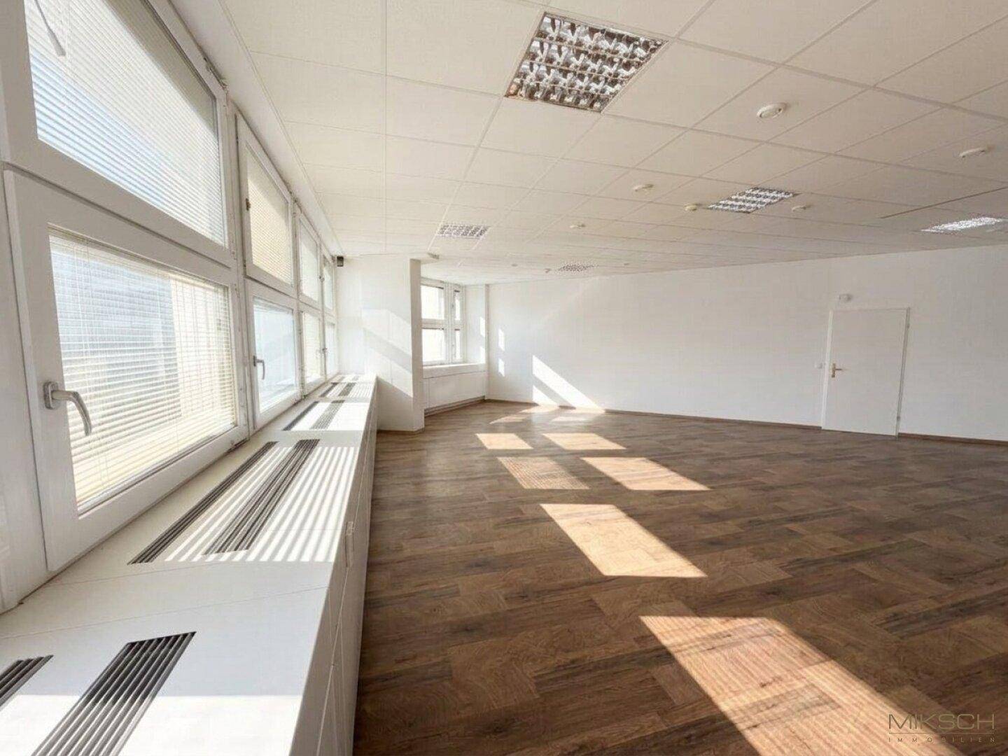 Helles Büro mit Holzboden, großen Fenstern und Jalousien, durchflutet von Sonnenlicht.