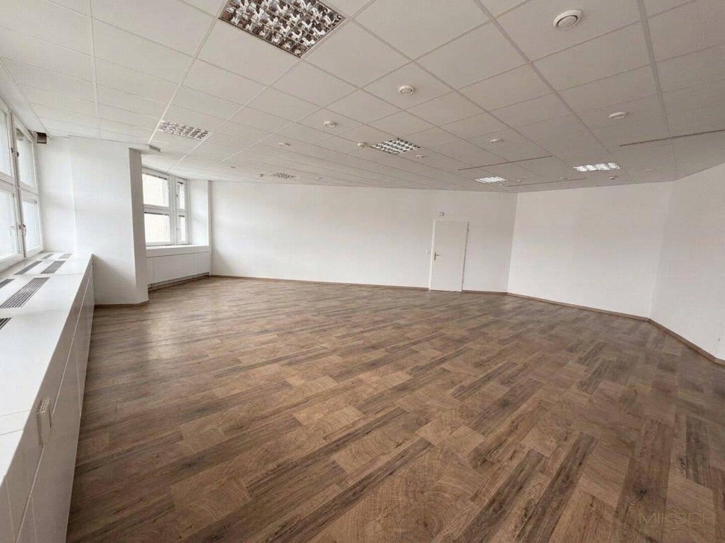 Leerer Büroraum mit Holzboden, weißen Wänden und Fenstern, die viel Licht hereinlassen.