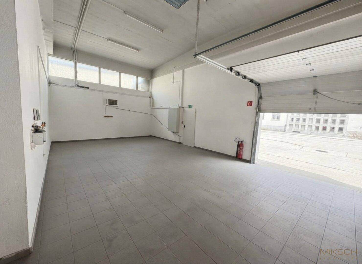 Leere Gewerbehalle mit Fliesenboden, Rolltor und Fenstern, die für ausreichend Tageslicht sorgen.