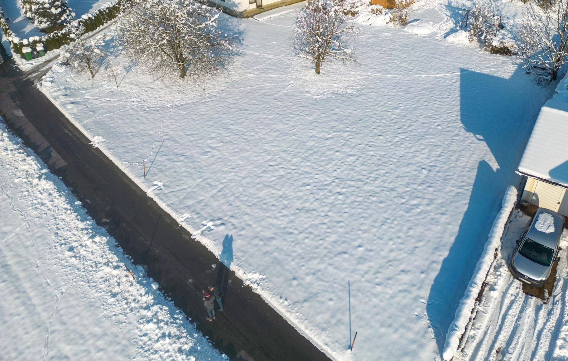Luftaufnahme eines schneebedeckten Grundstücks mit angrenzender Straße und Häusern in winterlicher Landschaft.