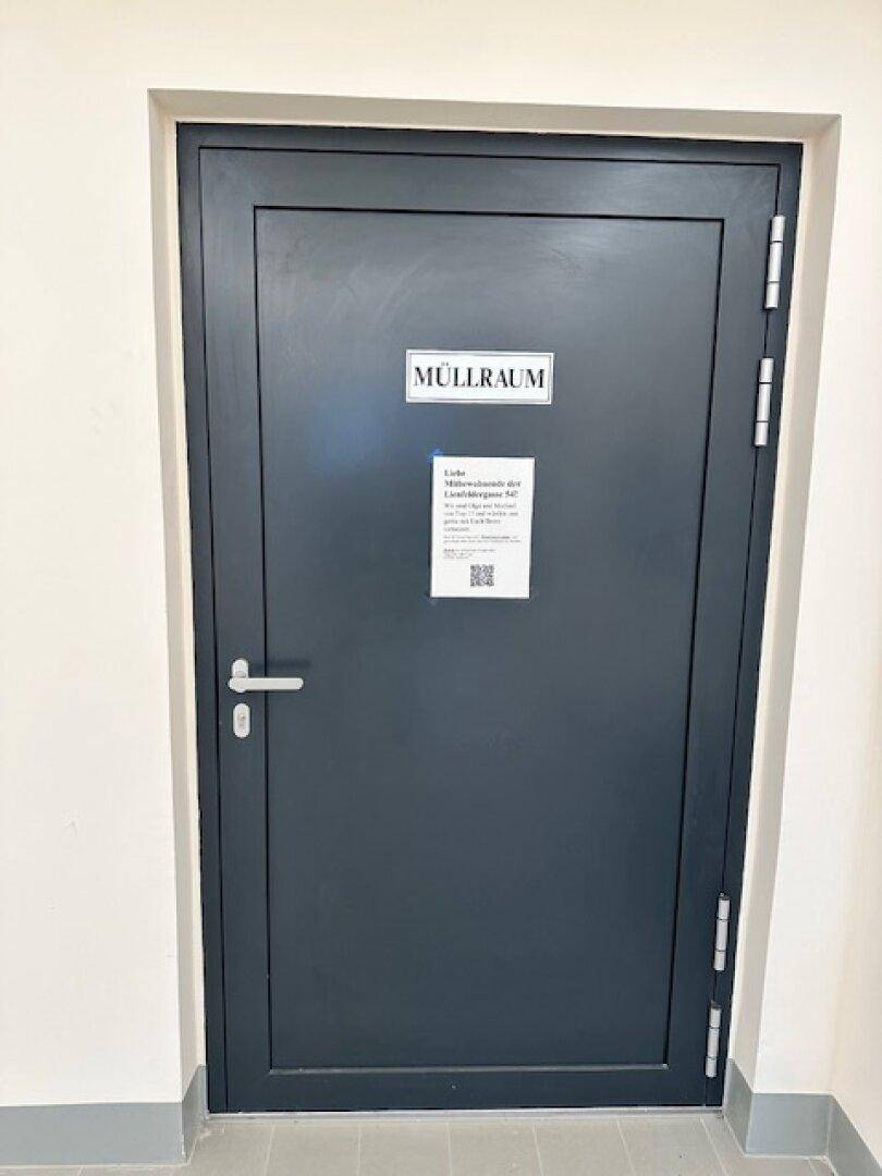 Dunkelgraue Tür zum Müllraum in einem Gemeinschaftsflur, mit Hinweisschildern versehen.