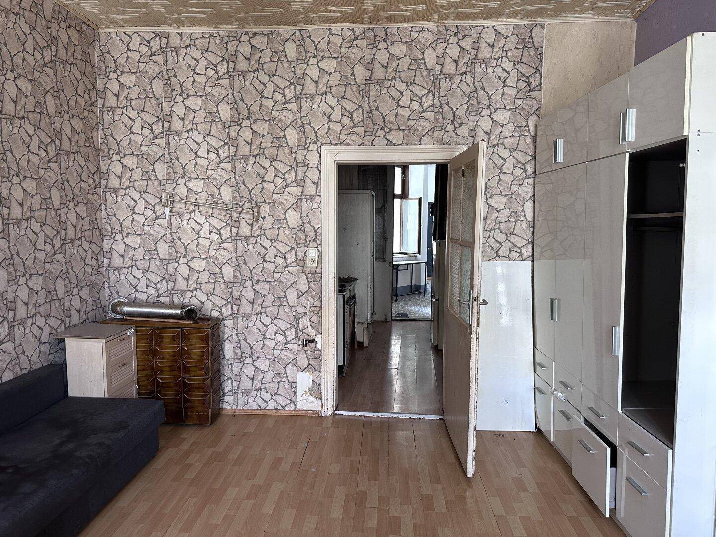 Wohnzimmer mit gemusterter Tapete, Holzboden und einem großen weißen Kleiderschrank.
