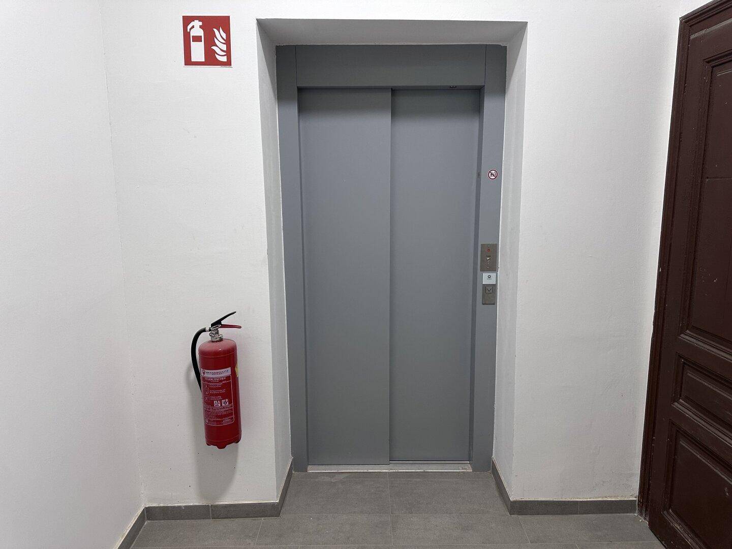 Moderner Aufzug in einem hellen Flur mit grauen Türen und einem Feuerlöscher.
