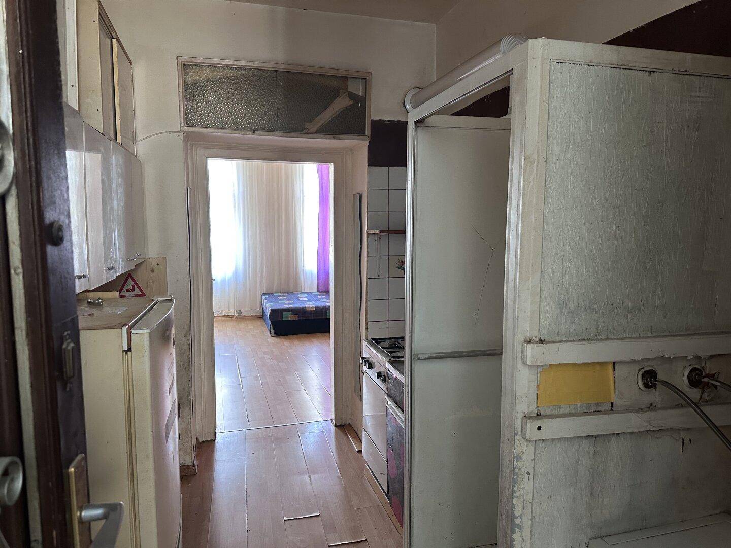 Blick durch einen Flur mit älteren Küchenelementen in ein angrenzendes Zimmer.