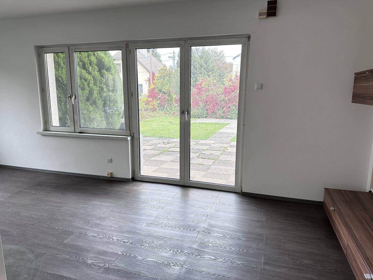 Heller Wohnbereich mit Laminatboden und breiten Fenstertüren, die den Blick in den grünen Garten freigeben.