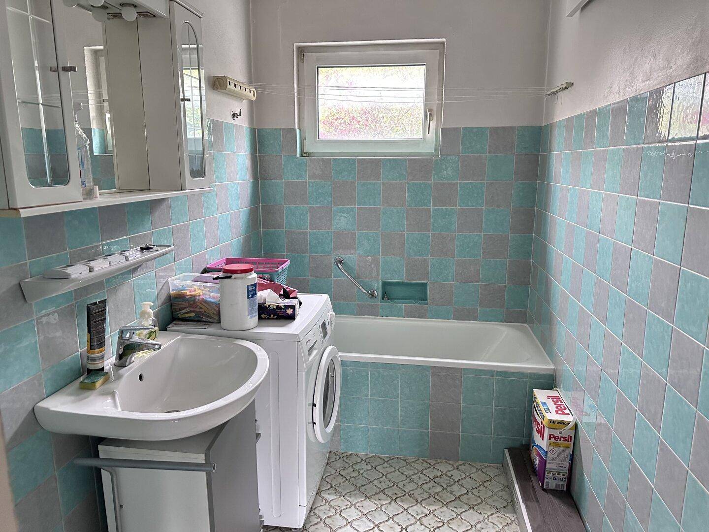 Badezimmer mit Badewanne, Waschmaschine und hellblauen sowie grauen Fliesen.