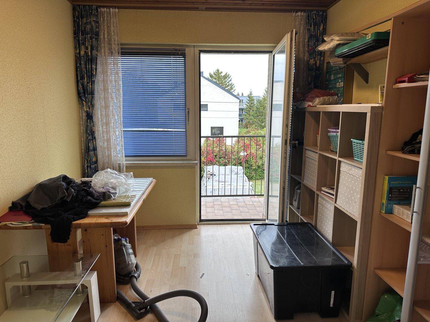 Helles Zimmer mit Laminatboden, Regalen und Zugang zu einem kleinen Balkon mit Blick ins Grüne.
