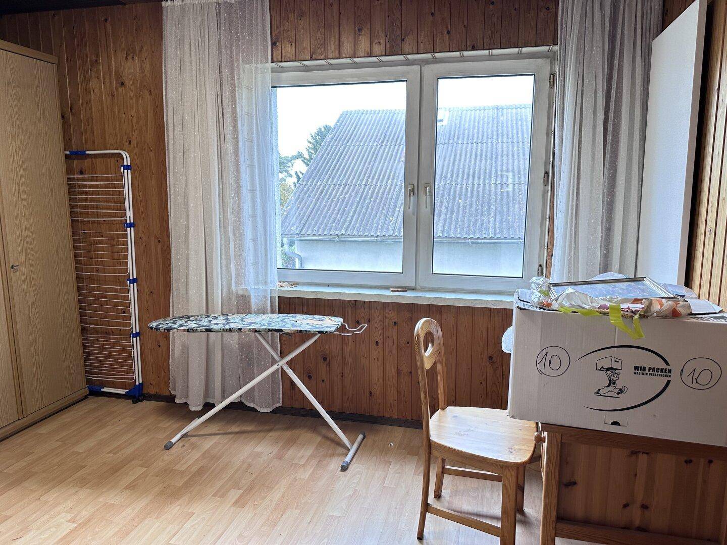 Helles Zimmer mit Holzvertäfelung, Laminatboden und großem Fenster mit Blick nach draußen.