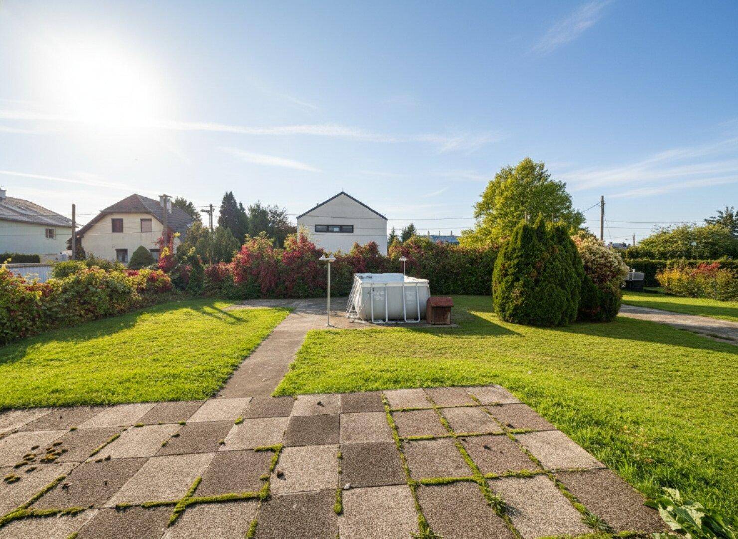 Weitläufiger Gartenbereich mit Rasen, gepflastertem Weg und einem Aufstellpool an einem sonnigen Tag.