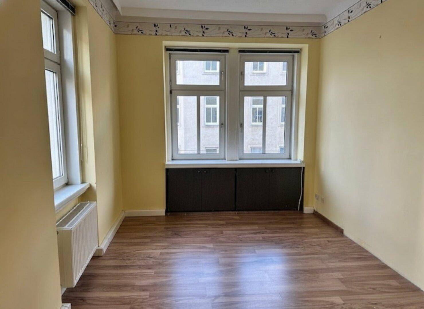 Helles Zimmer mit gelben Wänden, Laminatboden und praktischen Einbauschränken unter dem Fenster.