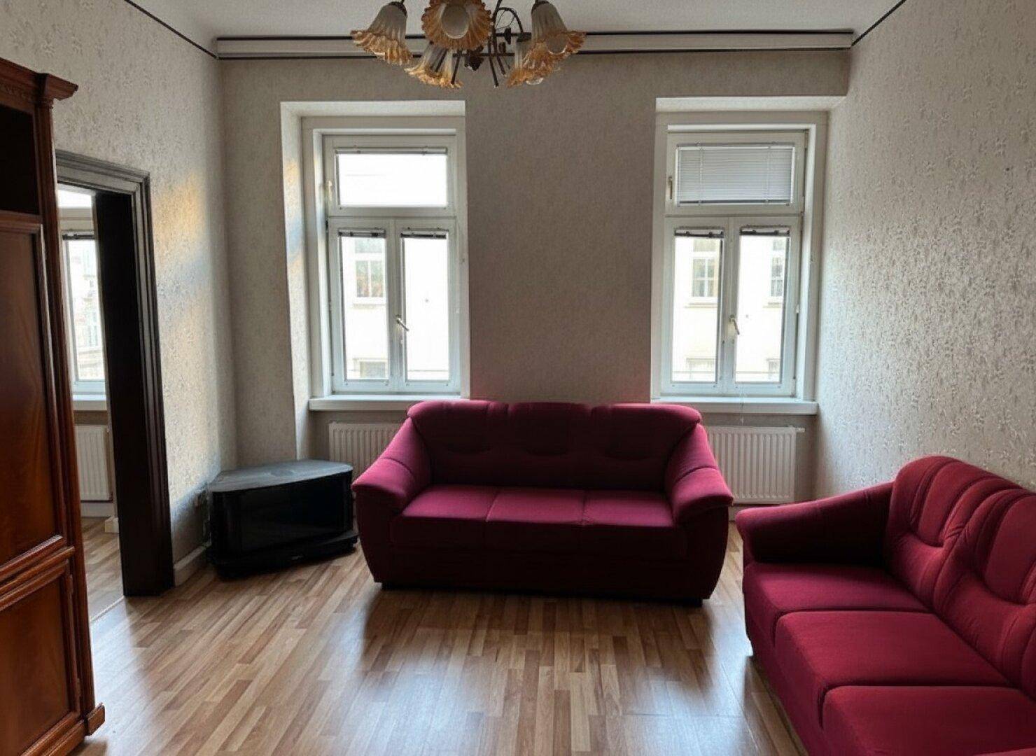Wohnzimmer mit Laminatboden, zwei Fenstern und bequemen roten Sofas.