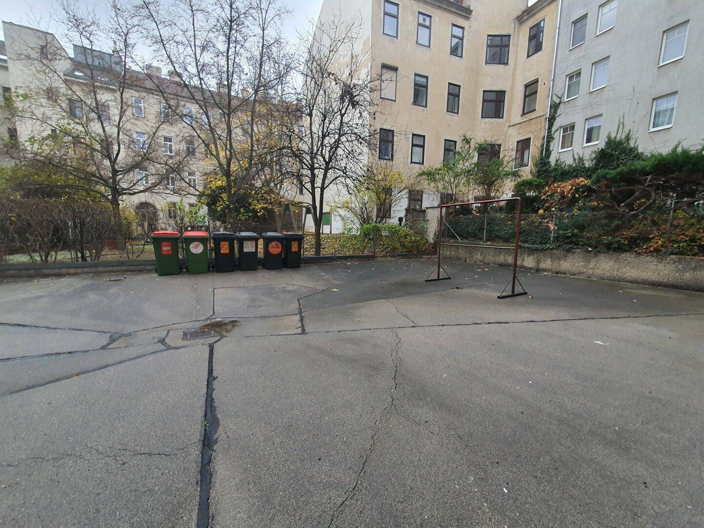 Hinterhof mit Mülltonnen, einem Fußballtor und Bäumen, umgeben von Gebäuden.