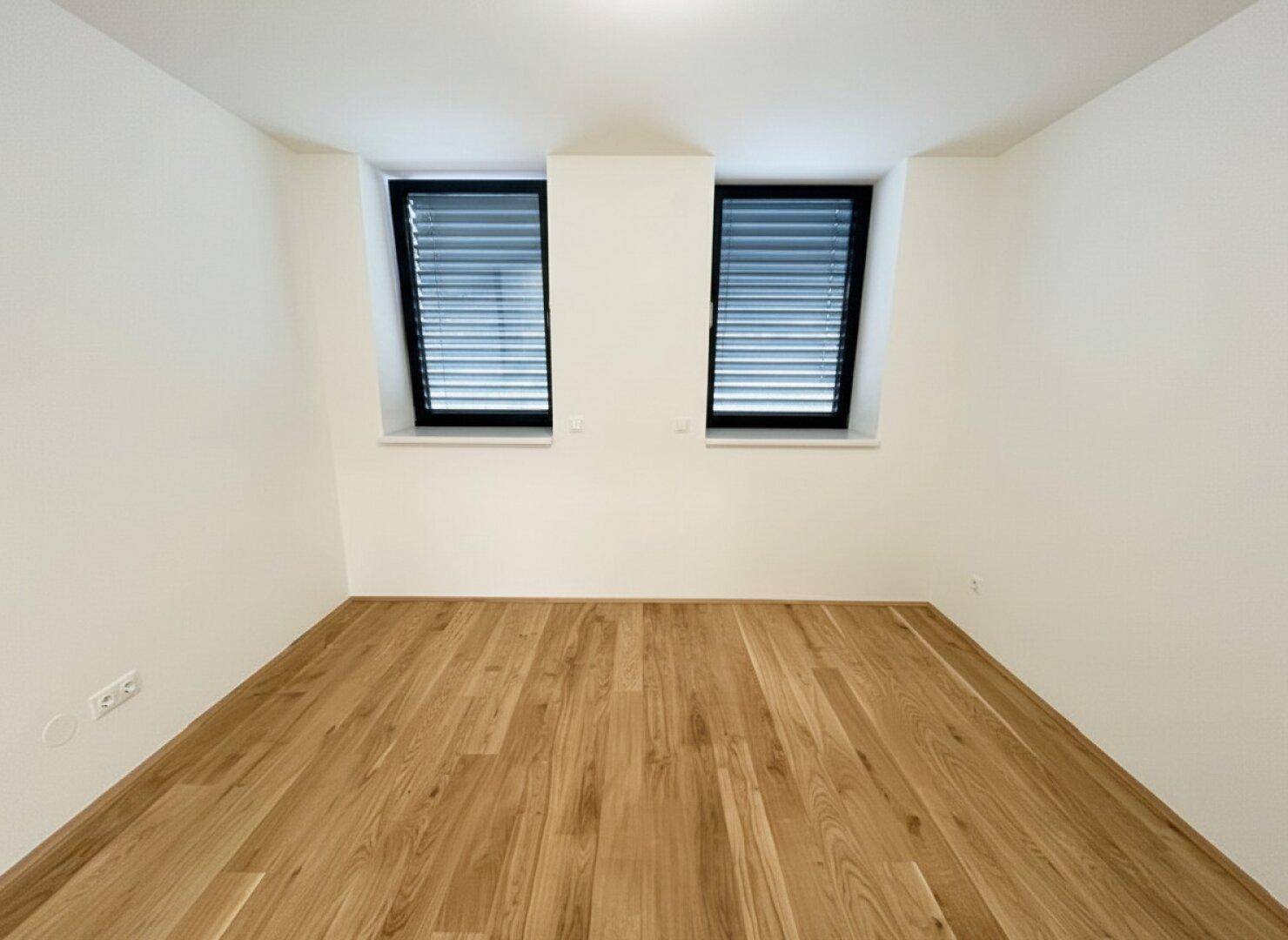 Leeres Schlafzimmer mit Holzboden und zwei Fenstern mit geschlossenen Jalousien für Privatsphäre.