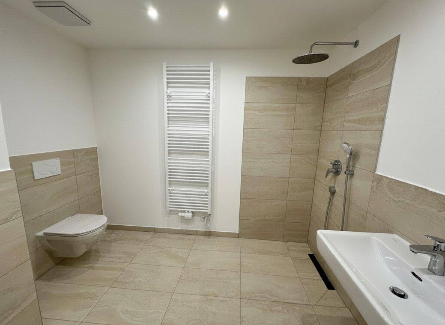 Komplettes Badezimmer mit beigefarbenen Fliesen, Toilette, ebenerdiger Dusche und Handtuchheizkörper.