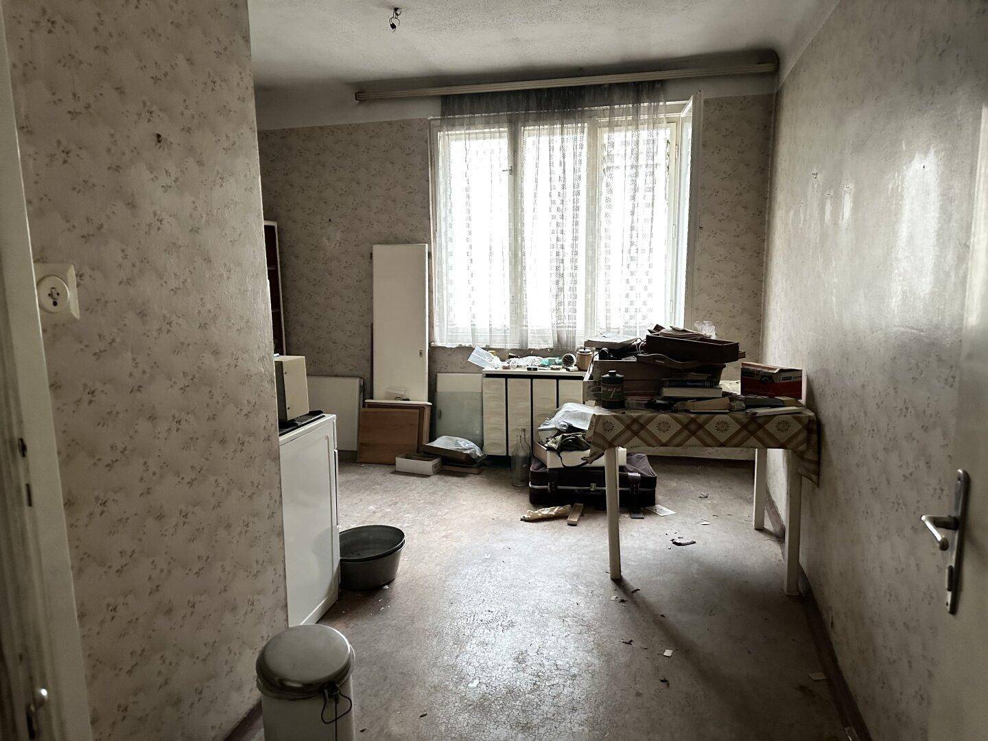 Unaufgeräumter Raum mit gemusterter Tapete, Fenster und verschiedenen Gegenständen.