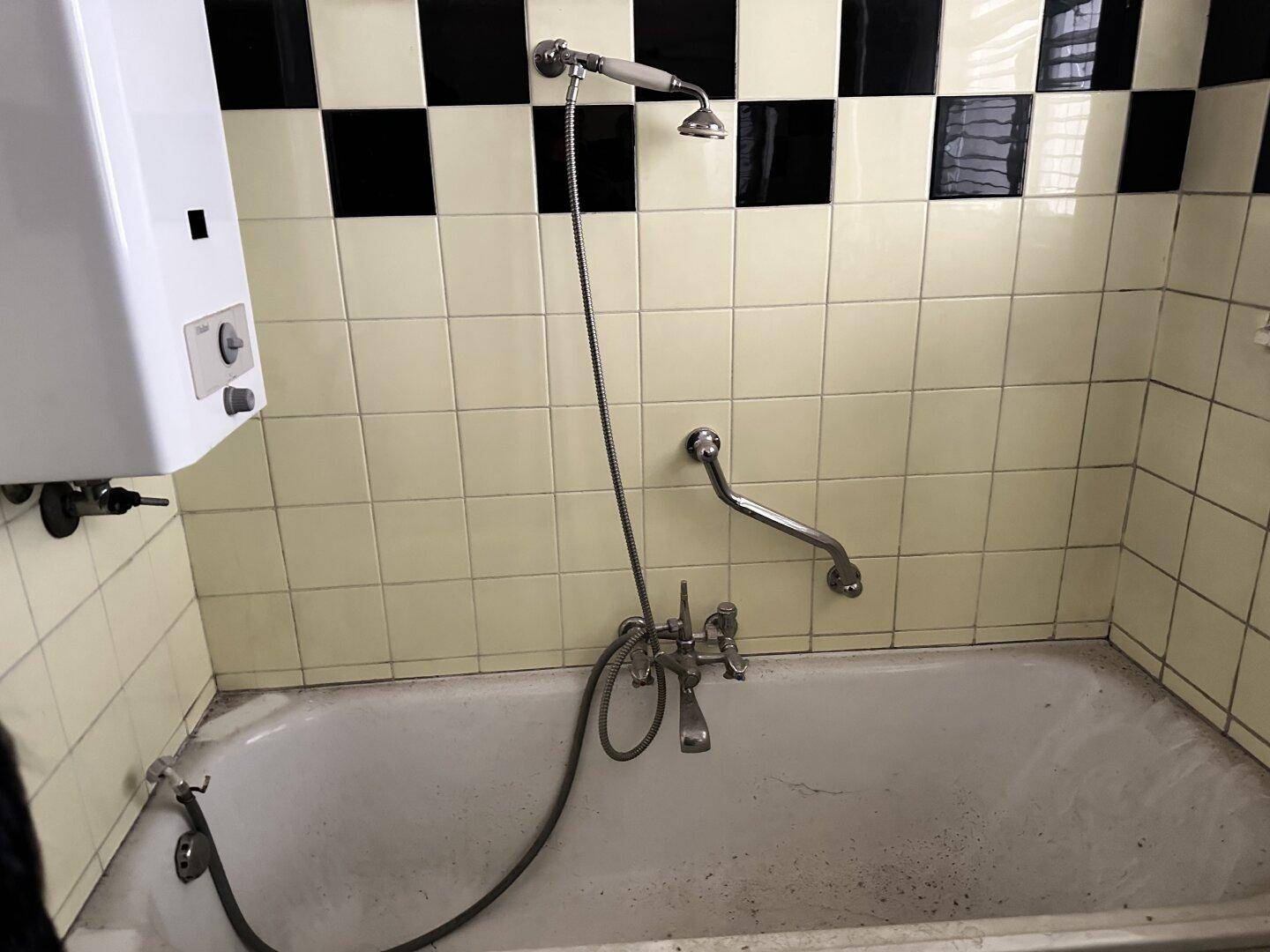 Badezimmer mit Badewanne, Duschkopf, Haltegriffen und gelb-schwarzen Fliesen im Retro-Stil.