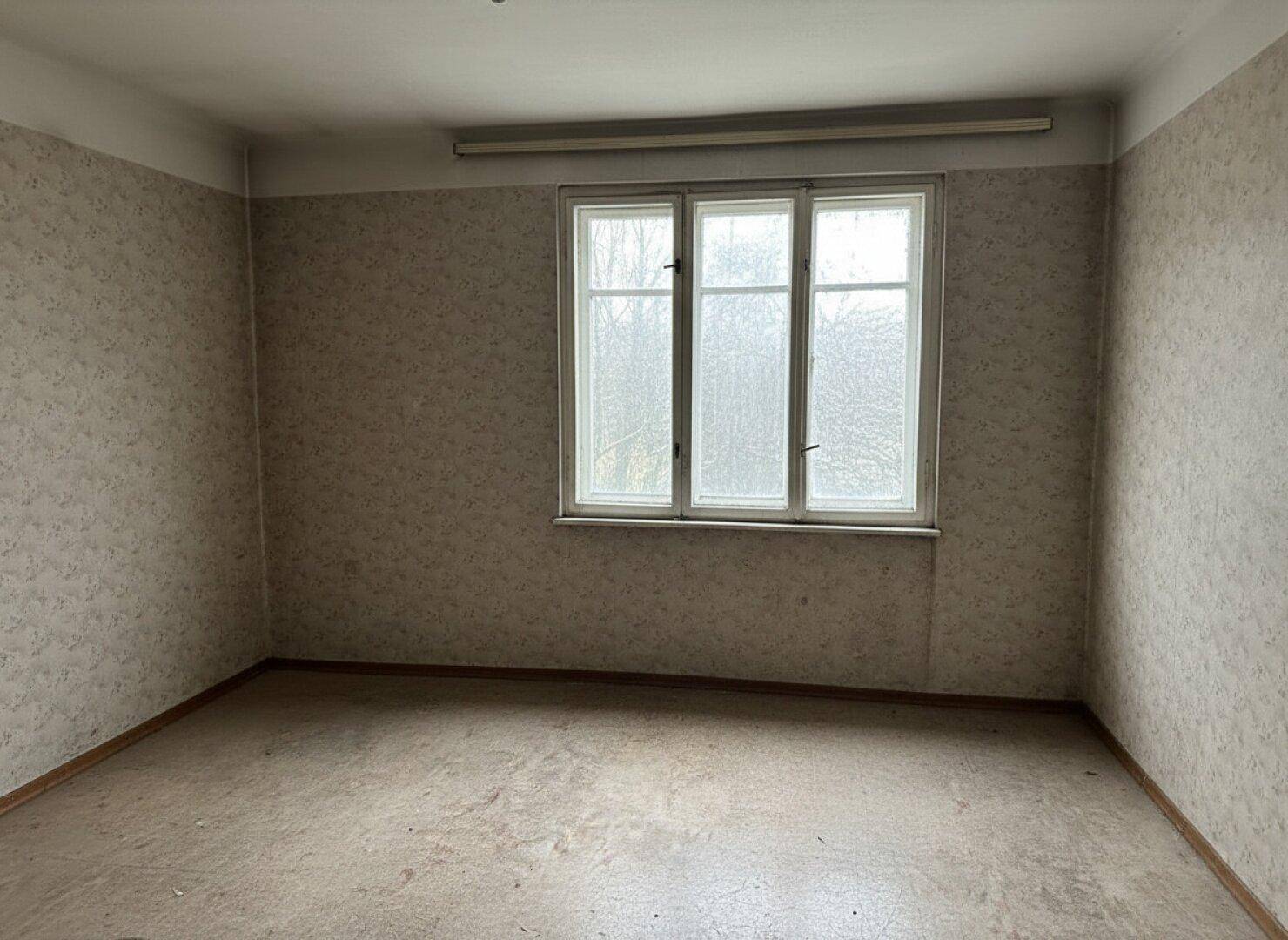 Leeres Zimmer mit gemusterter Tapete, altem Bodenbelag und einem Fenster.