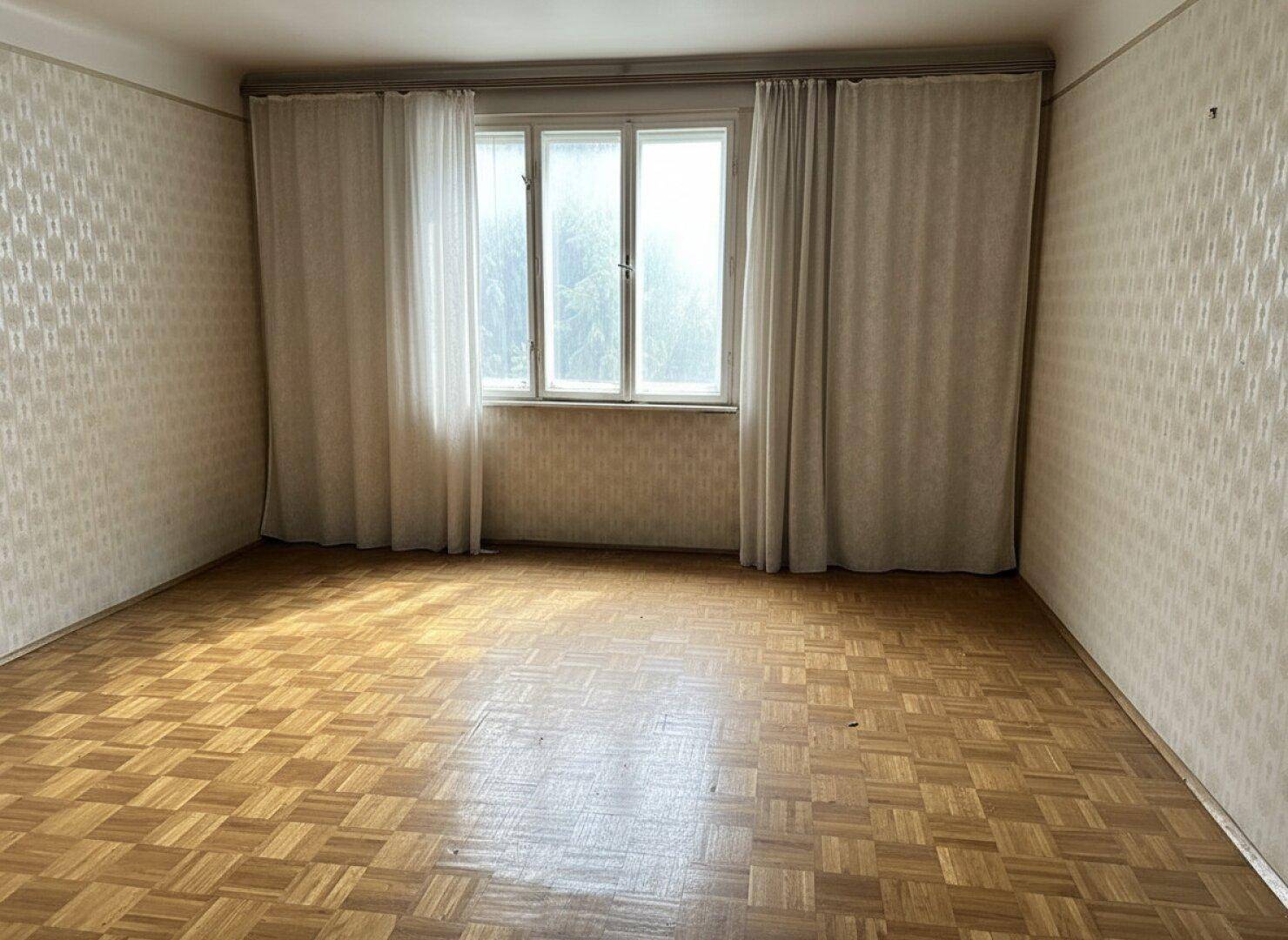 Geräumiges Zimmer mit Parkettboden, gemusterter Tapete und großem Fenster mit hellen Vorhängen.
