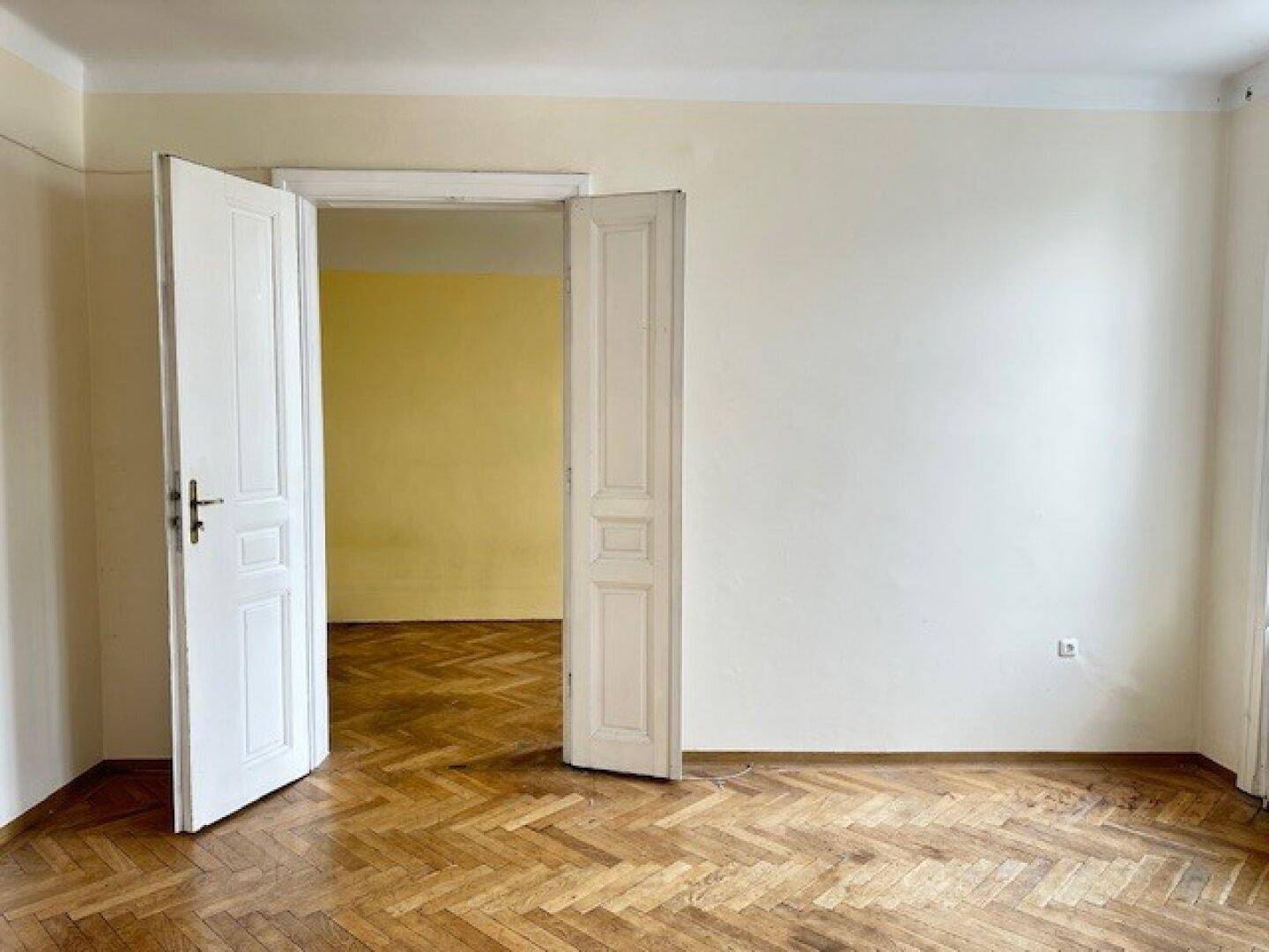 Geräumiges Zimmer mit Fischgrätparkett und offenen Doppeltüren zu einem angrenzenden Raum.