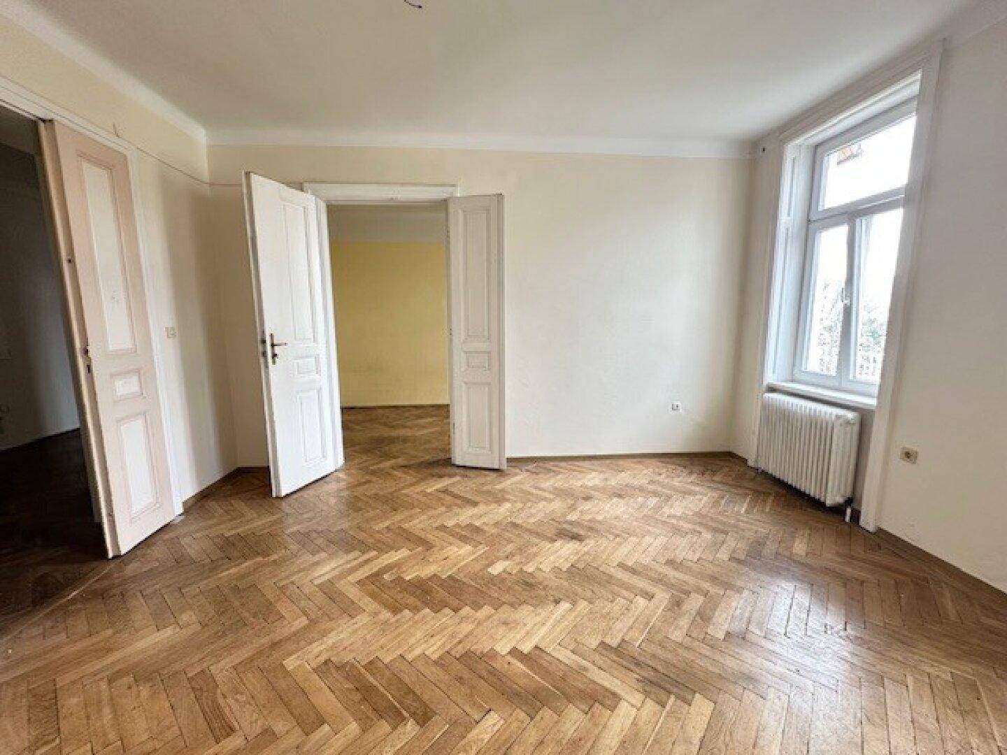 Heller Raum mit Fischgrätparkett, Doppeltüren und einem Fenster, das viel Tageslicht hereinlässt.