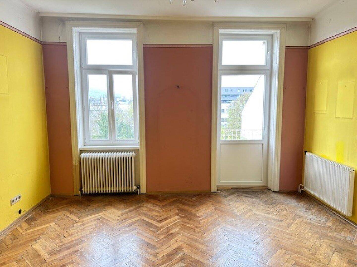 Helles Zimmer mit Fischgrätparkett, zwei Fenstern und farbigen Wänden, mit Zugang zum Balkon.