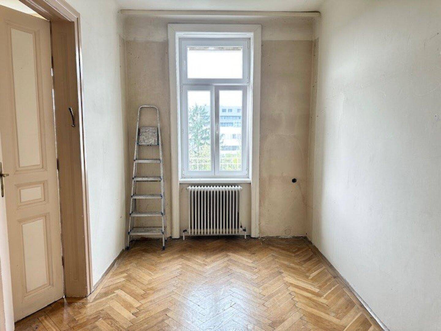 Schmaler Raum mit Fischgrätparkett, einem Fenster und einer Leiter, deutet auf Renovierungsarbeiten hin.