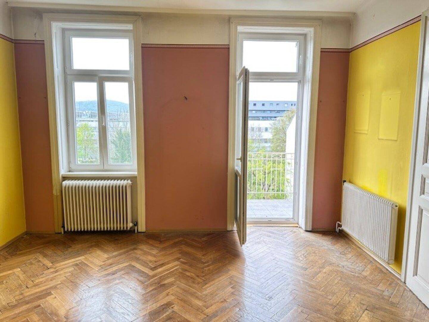 Heller Raum mit Fischgrätparkett, farbigen Wänden und offenem Zugang zu einem Balkon.