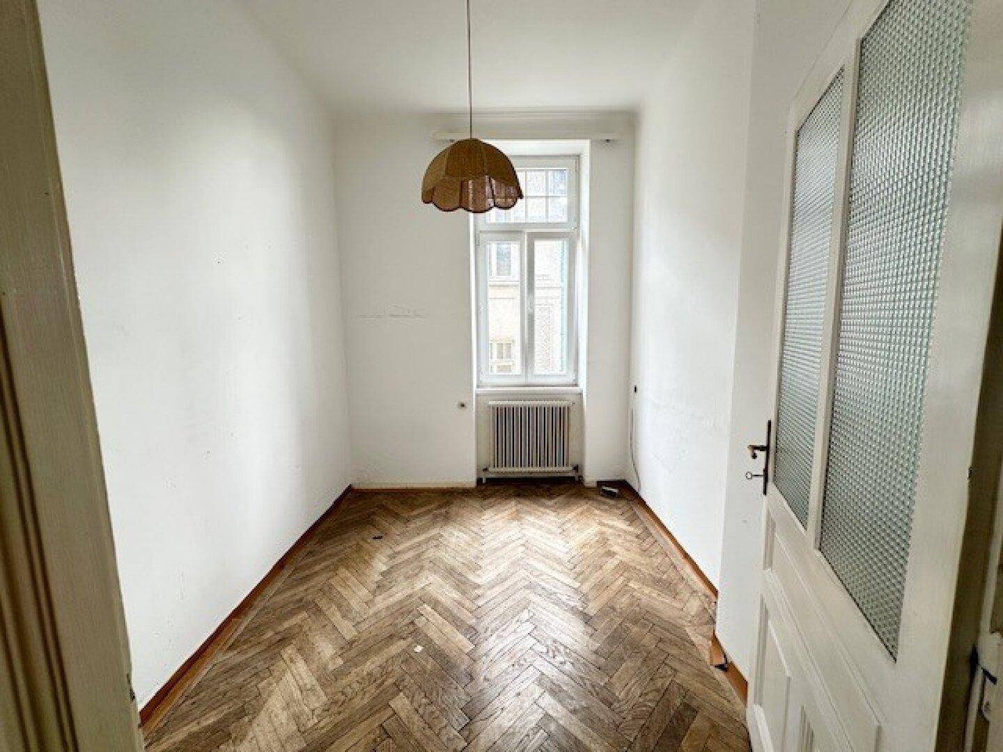 Schmaler Raum mit Fischgrätparkett, einem Fenster und einem dekorativen Deckenlicht.