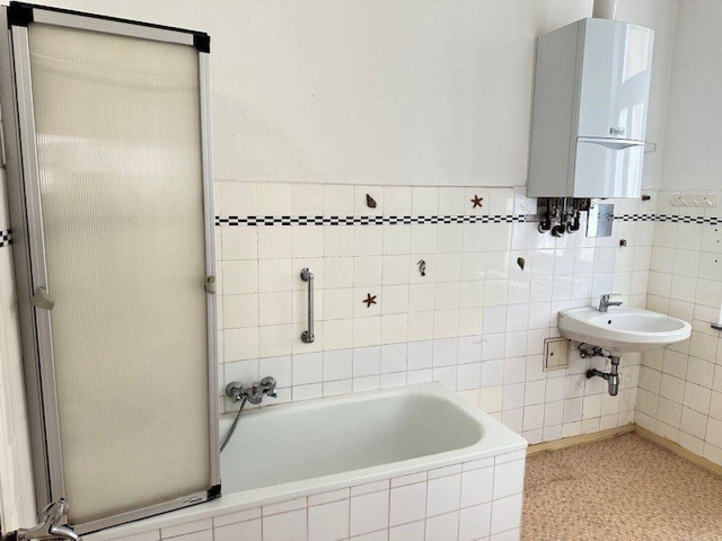 Badezimmer mit Badewanne, Duschabtrennung, Waschbecken und Wandheizkessel, in einfachem Zustand.