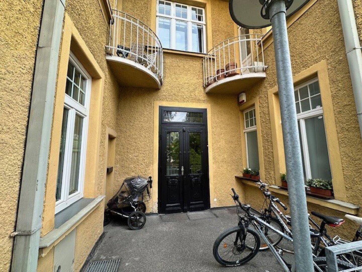 Eingangsbereich des gelben Gebäudes mit schwarzer Doppeltür, Fenstern und abgestellten Fahrrädern.