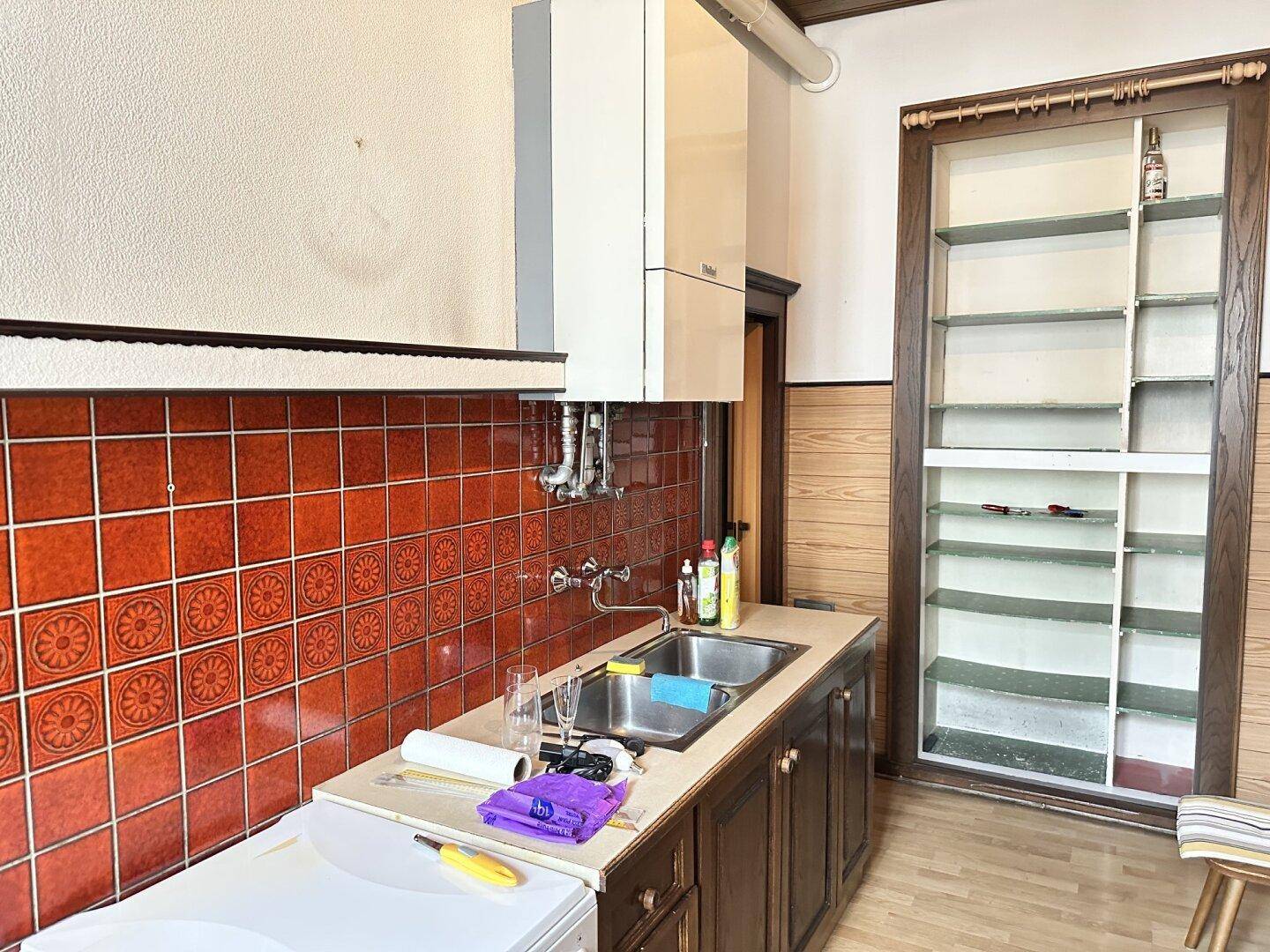 Küchenzeile mit Spülbecken, roten Fliesen und Boiler, daneben ein offenes Regal.