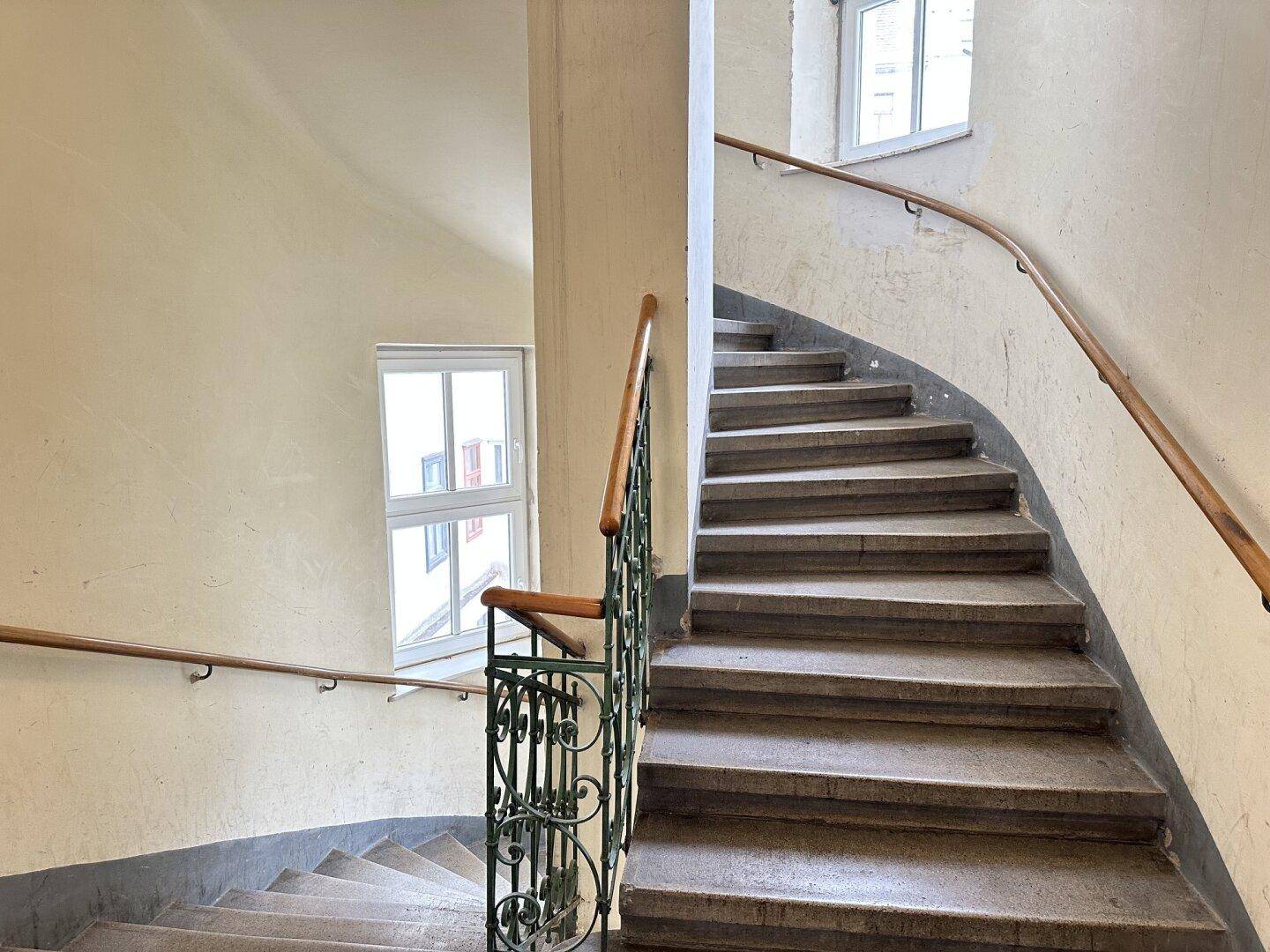 Historisches Treppenhaus mit Steinstufen, schmiedeeisernem Geländer und Fenstern.