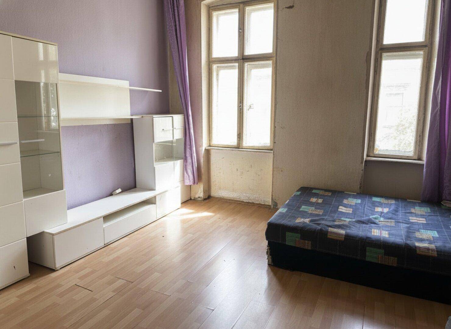 Helles Schlafzimmer mit Holzboden, lila Akzentwand und viel Tageslicht durch zwei Fenster.