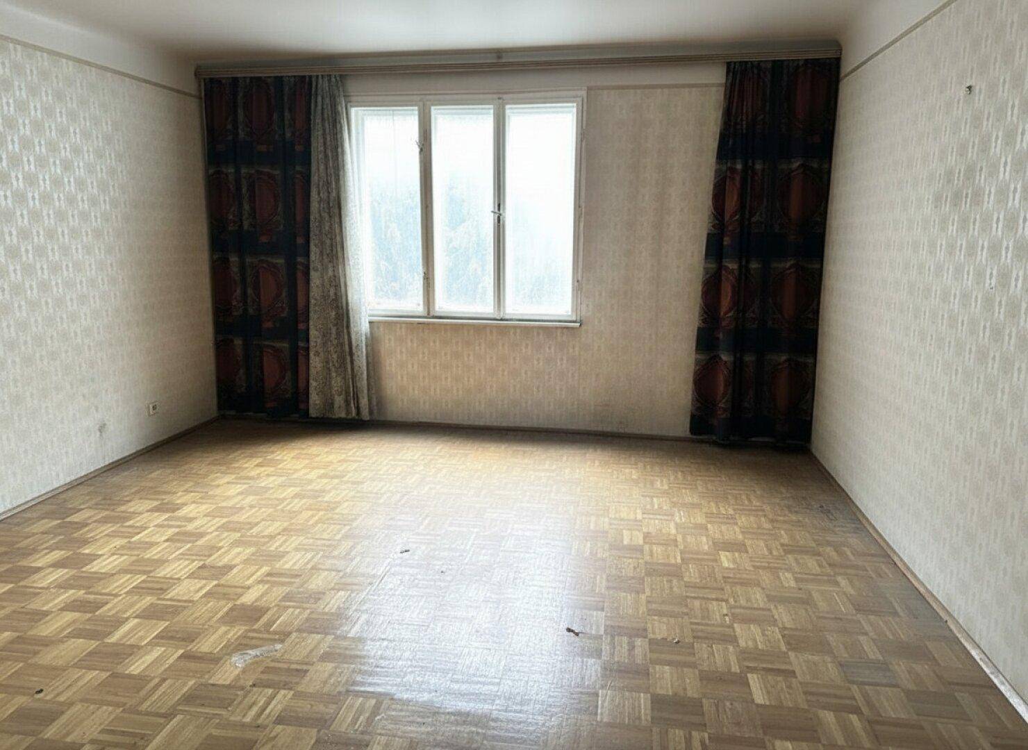 Leeres Zimmer mit Parkettboden, gemusterter Tapete und dunklen Vorhängen am Fenster.