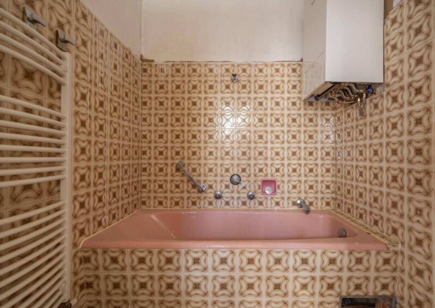 Badezimmer mit gemusterten braunen Fliesen, einer rosa Badewanne und einem Handtuchheizkörper.