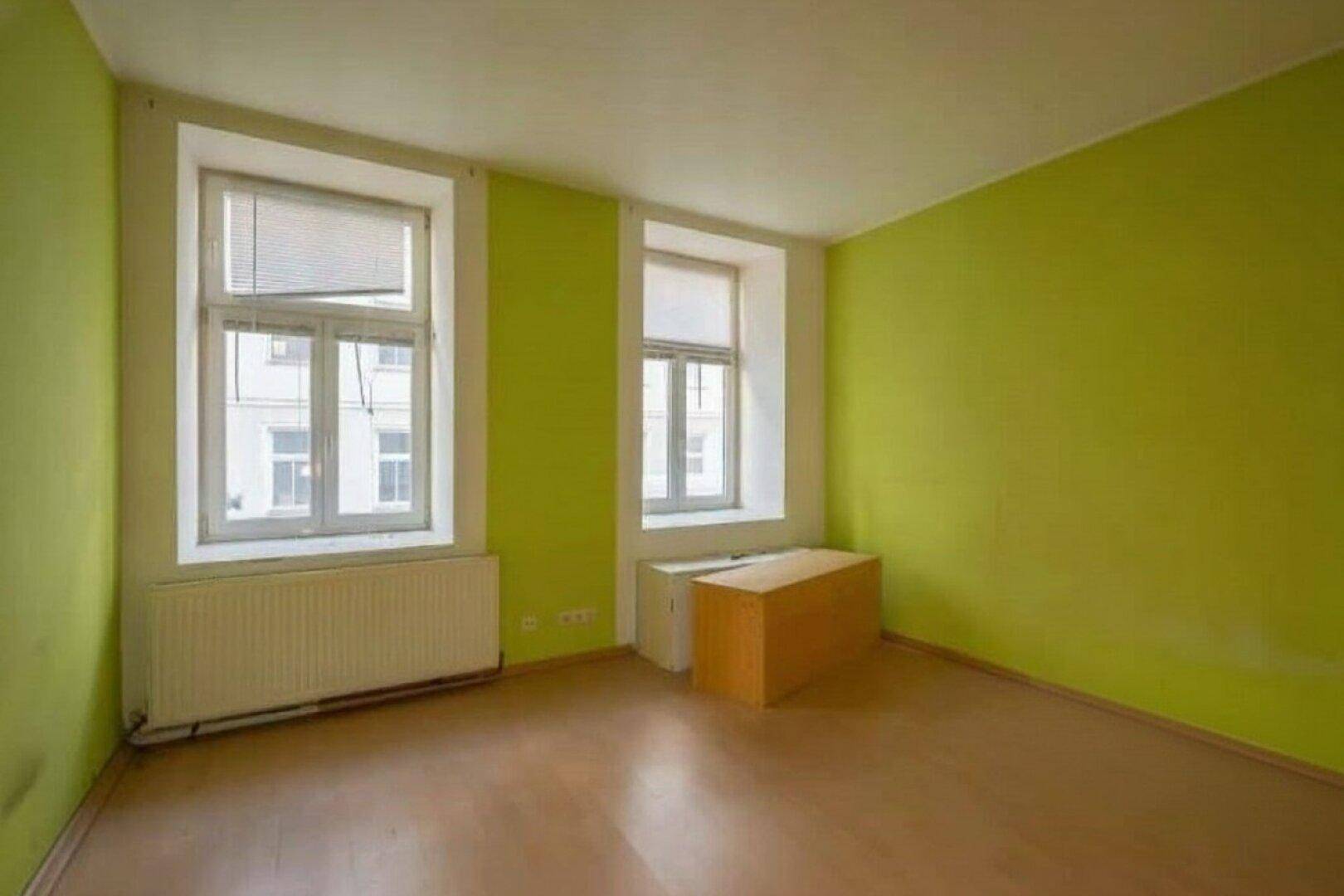 Leerer Raum mit auffällig grünen Wänden, zwei Fenstern und hellem Holzfußboden.