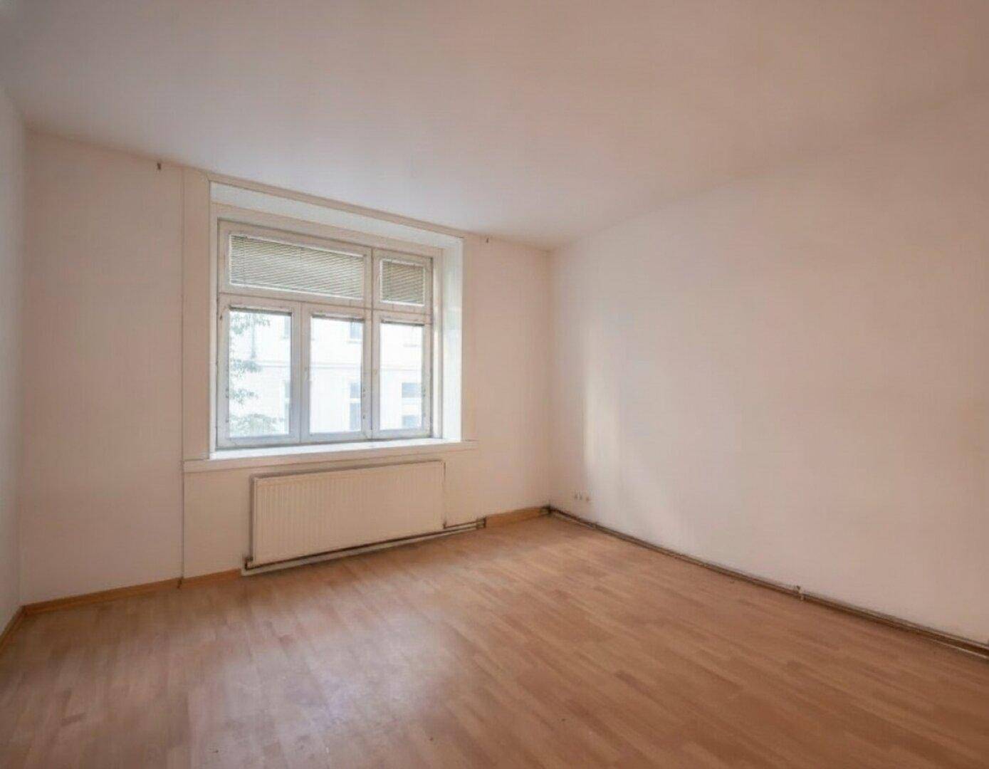 Heller, leerer Raum mit weiß gestrichenen Wänden, Holzfußboden und einem Fenster mit Heizkörper.