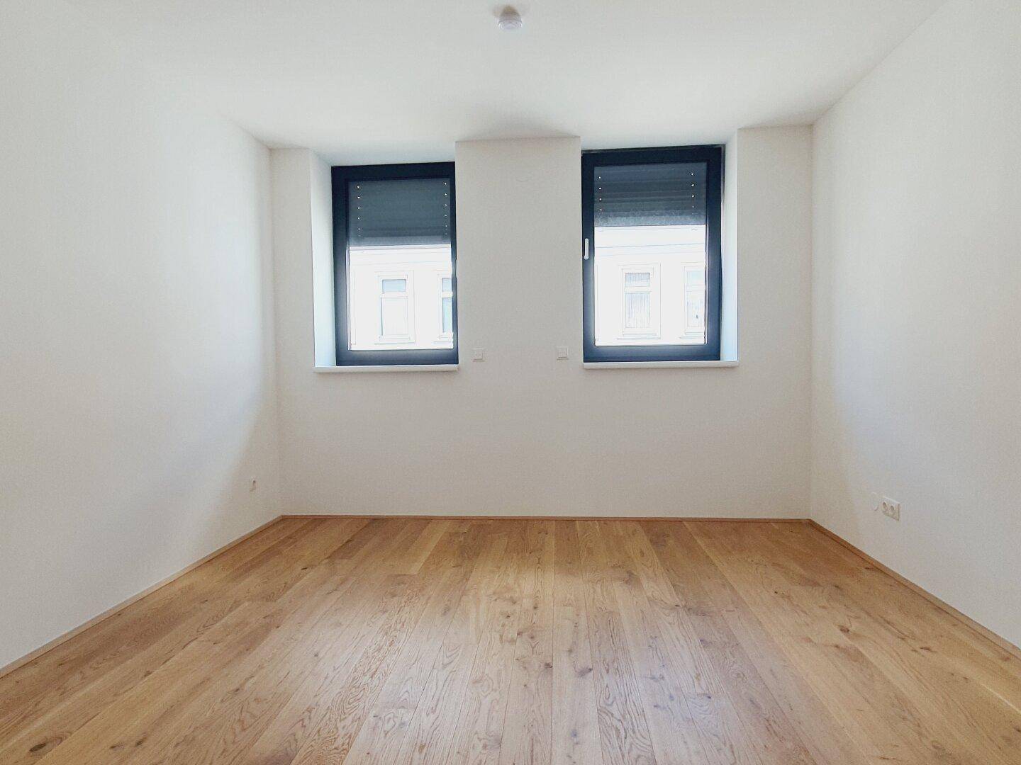 Helles Schlafzimmer mit Holzboden und zwei Fenstern, die einen Blick nach draußen ermöglichen.